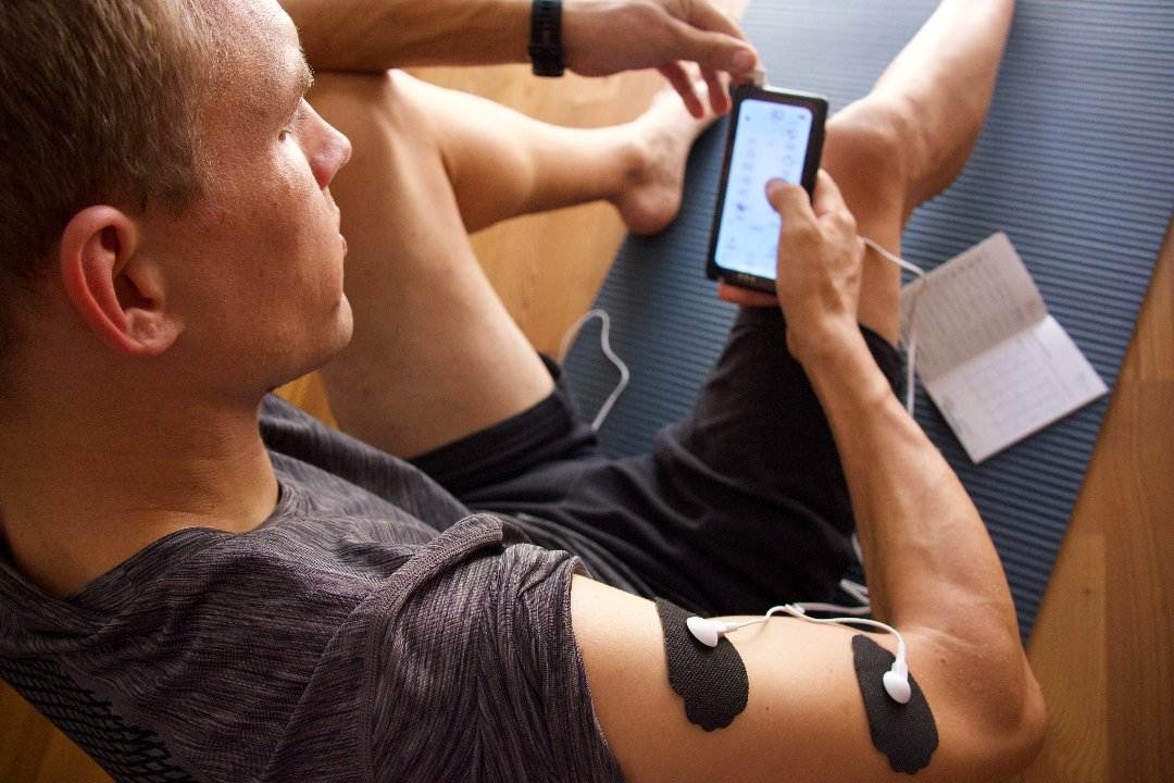 Person benutzt das OrthoMechanik TENS EMS Elektro-Stimulationsgerät zur Muskelentspannung. Elektroden-Pads sind am Oberarm angebracht, während die Person das Gerät bedient und die Intensität oder den Modus anpasst. Perfekt für gezielte Muskelbehandlungen nach dem Sport oder zur Schmerzlinderung im Alltag.