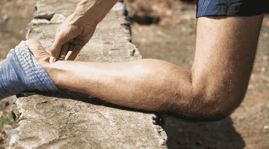 Nahaufnahme eines Sportlers, der seine Achillessehne massiert, während sein Fuß auf einer Mauer ruht. Er trägt eine blaue Socke und kurze Sportkleidung