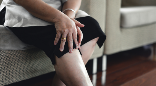 Ältere Frau sitzt auf dem Sofa und hält sich schmerzhaft das Knie – Anzeichen für Gelenkschmerzen, Arthrose oder Muskelverspannungen im Bein