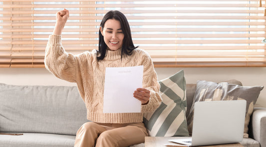 Glückliche Frau auf Sofa hebt jubelnd eine Rückerstattungsbescheinigung