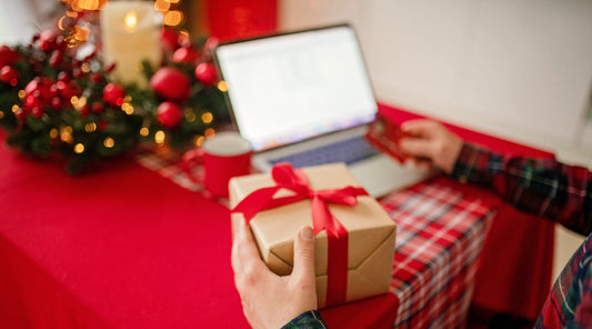 Mann hält Geschenkbox vor Laptop mit Weihnachtsdeko.