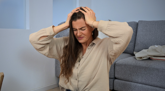 Eine Frau mit langen Haaren sitzt vor einem Sofa und hält sich mit schmerzverzerrtem Gesicht den Kopf, was auf Kopfschmerzen oder Migräne hindeutet. Die Szene spielt in einem wohnlichen Umfeld.