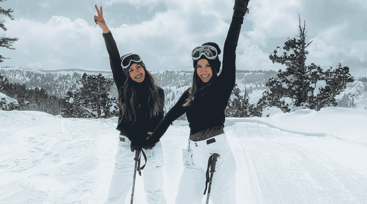 Zwei Frauen posieren fröhlich auf einer verschneiten Piste vor winterlicher Berglandschaft.