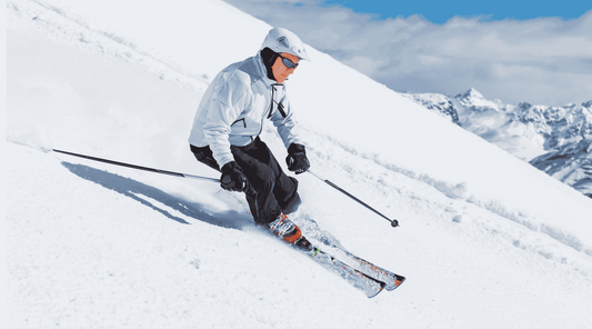 Ein Skifahrer in weißer Winterausrüstung fährt eine schneebedeckte Piste hinunter. Im Hintergrund sind schneebedeckte Berge und ein blauer Himmel zu sehen. Die Szene vermittelt Geschwindigkeit und Kontrolle beim Skifahren.