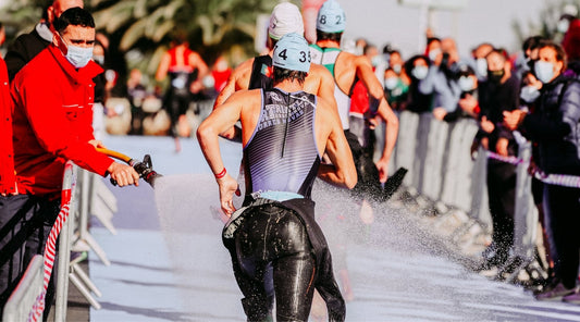Triathleten beim Wechsel nach dem Schwimmen – Wettkampfszene mit Wasserdusche und jubelnden Zuschauern