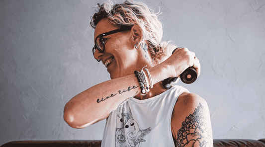 Eine lächelnde Frau mit Brille und sichtbaren Tattoos nutzt eine Massagepistole, um Verspannungen im Nacken- und Schulterbereich zu lösen. Sie trägt ein ärmelloses Oberteil, und der Hintergrund ist neutral gestaltet.